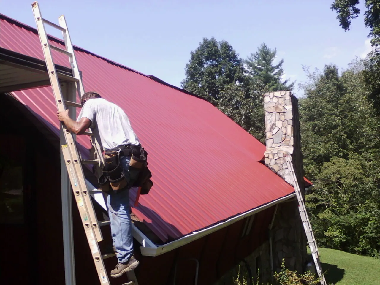 Roofer on a ladder beside a red metal roof and stone chimney — Soffit & Fascia Repair by Roofing Brothers in Elk Point